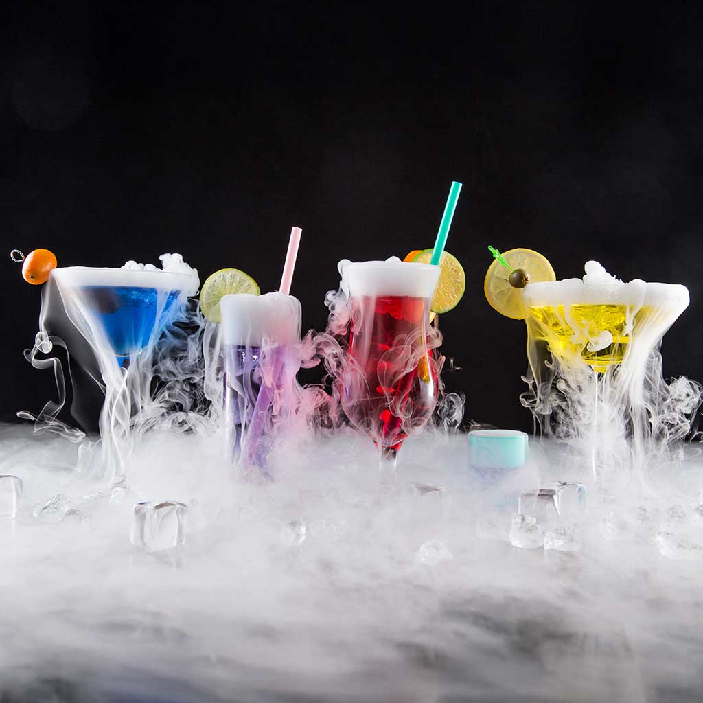 Dry ice creating a fog effect in a grouping of cocktails on a bar top.
